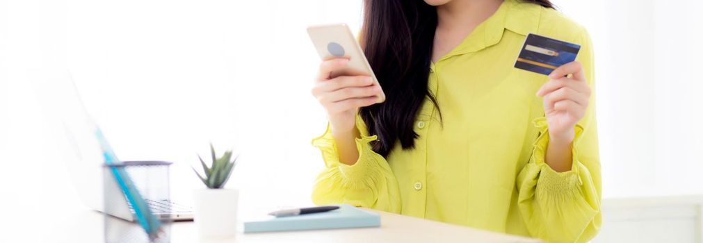 Young asian business woman using smart phone and holding credit card while online shopping and payment online with laptop computer on desk at home, female holding debit card, communication concept.