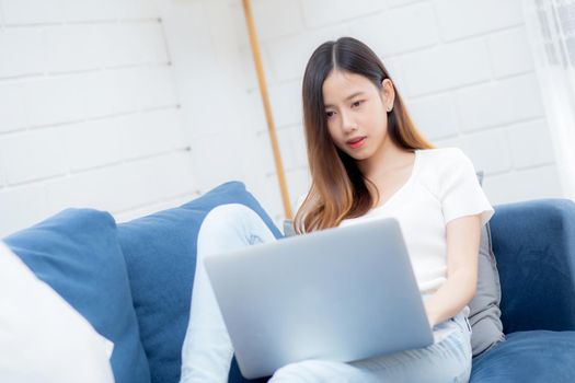 Young asian business woman smile and work from home with laptop computer online to internet on sofa in living room, freelance girl using notebook on couch with comfort, new normal, lifestyle concept.