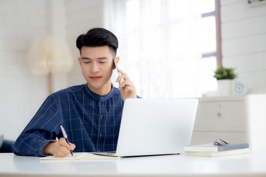 Young asian man talking phone and work from home with laptop computer, freelance male writing on notebook and speak on smartphone, stay home, domestic life, business and communication concept.