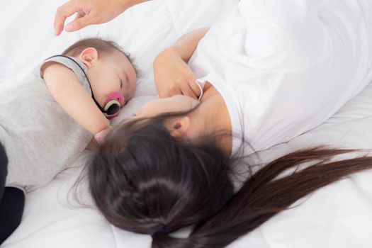 Young asian mother touch head of little baby girl with tender on bed in the bedroom, mom love newborn and care, woman with expression with child together, parent and daughter, family concept.