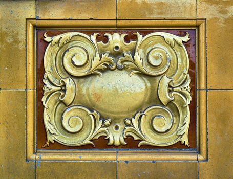 A close up look at the detail of some warm yellow and gold victorian tiling that shows some age and weathering. The tiles have a pattern of scrolls and plants.