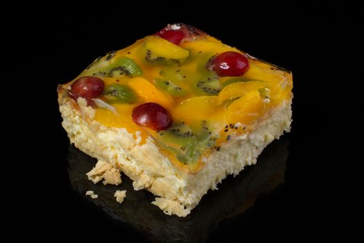 A piece of a cake covered with yellow jelly with fruit close-up on a dark black background