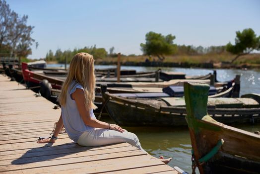 Blonde girl sitting on a jetty, small boats, no shoes, lifestyle
