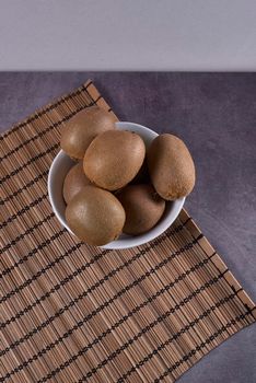 Several kiwis in a bowl on a reed tablecloth, stone floor, white and brown.