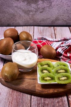 Glass with yoghurt and broken kiwis on a plate. Wooden floor and wooden board, whole kiwis, white and red kitchen towel.