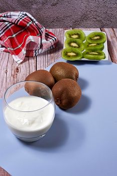 Glass with yoghurt and broken kiwis on a plate. Wooden floor and wooden board, whole kiwis, white and red kitchen towel, blue mat.