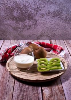 Glass with yoghurt and broken kiwis on a wooden plate. Wooden floor, whole kiwis, white and red kitchen towel, stone wall.