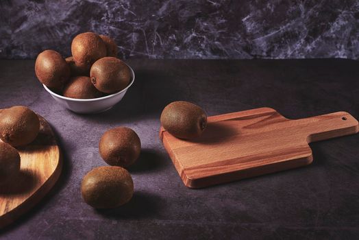 Three kiwis on a wooden board and one in a bowl. Dark stone floor and marble wall, white bowl.