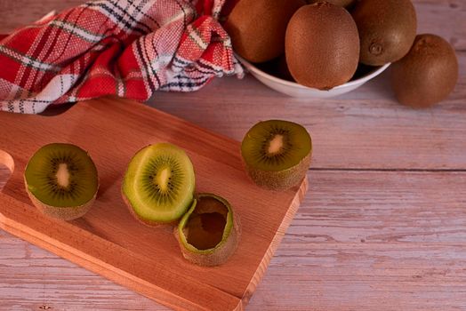 kiwis in white bowl and wooden board, wooden floor, kitchen towel, checkered, red, warm tones, split and whole kiwis, skin. dark food