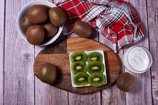 Plate of cut kiwis and several whole kiwis on wood. Wooden board, red and white kitchen towel, squares, white bowl, top view.