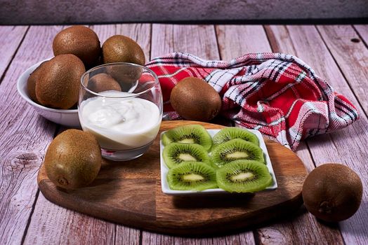 Yoghurt glass and plate of cut kiwis and several whole ones on wood. Wooden board, red and white tea towel, squares, white bowl, front view.