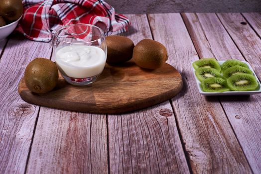 Yoghurt glass and plate of cut kiwis and several whole ones on wood. Wooden board, red and white tea towel, squares, white bowl, front view.
