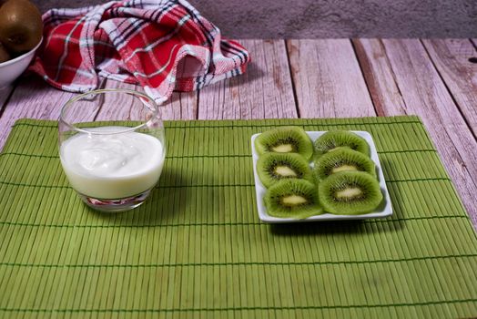 Yoghurt cup and plate of kiwis cut into rectangular plates, green wooden tablecloth, red and white kitchen towel, pictures, front view