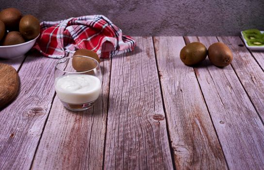 Glass of yoghurt with kiwis. Wooden floor, red and white kitchen towel, pictures, stone wall.