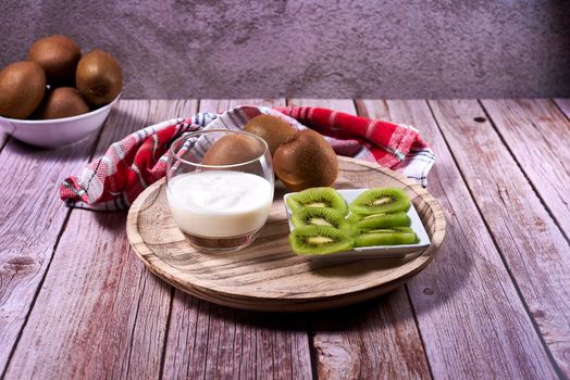 Yoghurt glass with kiwis on wooden plate. Wooden floor, red and white kitchen towel, chequered, zenithal view, kiwi cut on rectangular plate.