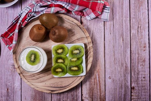 Yoghurt glass with kiwis on wooden plate. Wooden floor, red and white kitchen towel, chequered, zenithal view, kiwi cut on rectangular plate.