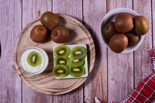 Yoghurt glass with kiwis on wooden plate. Wooden floor, red and white kitchen towel, chequered, zenithal view, kiwi cut on rectangular plate.