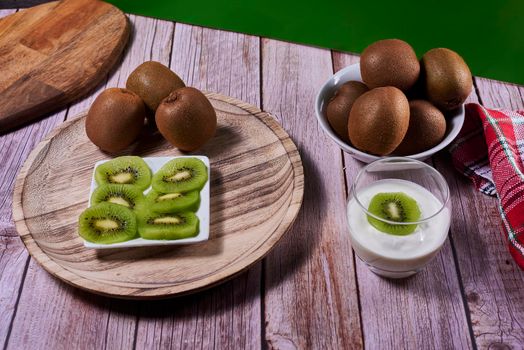 Yoghurt glass with kiwis on wooden plate. Wooden floor, red and white kitchen towel, chequered, front view, kiwi cut on rectangular plate.