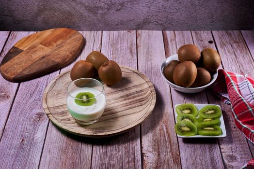 Yoghurt glass with kiwis on wooden plate. Wooden floor, red and white kitchen towel, chequered, front view, kiwi cut on rectangular plate.