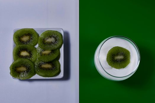 Yoghurt glass with kiwi fruit and plate with kiwi fruit cut on green and white background, rectangles, horizontal, symmetry