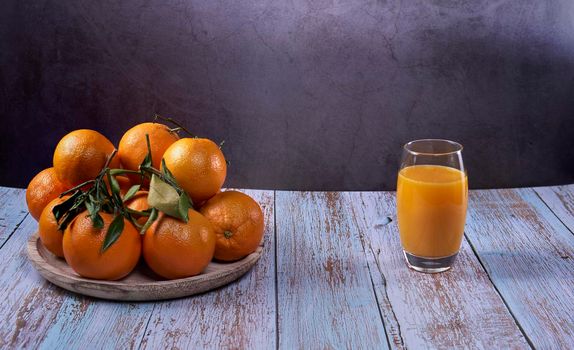 Large glass of orange juice and several oranges on wooden plate on wooden floor and marble background