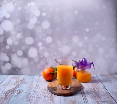 Glass of orange juice with glass juicer on wooden table, various oranges on wooden floor and bright background