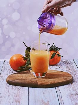 Hand pouring orange juice into glass with glass squeezer, wooden table, wooden floor bright background