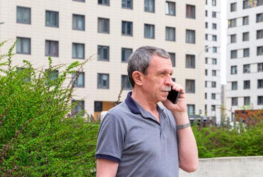 Adult caucasian senior man in a T-shirt talking on a smartphone in the city in spring or summer against the background of buildings. Selective focus.