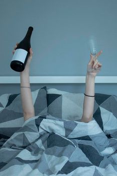 Hands of a girl from under a blanket holding a bottle of wine and a glass - grey sheets.