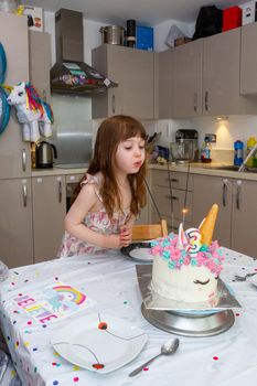 A Cute, brown-haired baby girl in a kitchen blowing candles of a birthday cake decorated as an unicorn with a number three candle