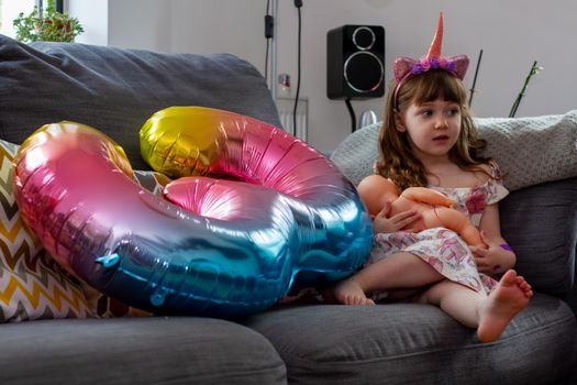A Cute Baby Girl Sitting On A Sofa With A Colourful Balloon And A Doll