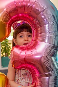 Portrait of a cute, brown-haired girl holding a colourful number three balloon and smiling at camera