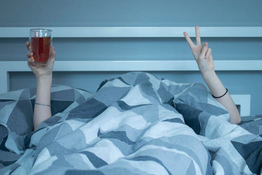 Hands of a girl from under a blanket holding a cup of tea with a double bottom, showing a pease sign with her fingers