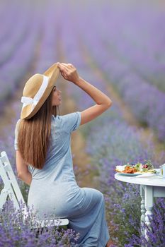 Back view of young woman wearing straw hat and dress sitting at table served with croissants, orange juice and plate. Attractive girl sitting in lavender field, looking aside, outdoors restaurant.