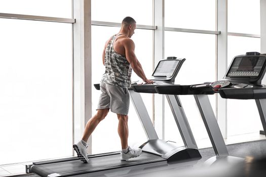 Muscular athletic bodybuilder fitness model running treadmill gym near big window jogging stationary machine do cardio training Weight loss Healthy lifestyle concept. Back view rear full length
