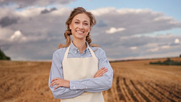 Woman farmer apron standing farmland smiling Female agronomist specialist farming agribusiness Happy positive caucasian worker agricultural field Pretty girl arms crossed cloudy sky background