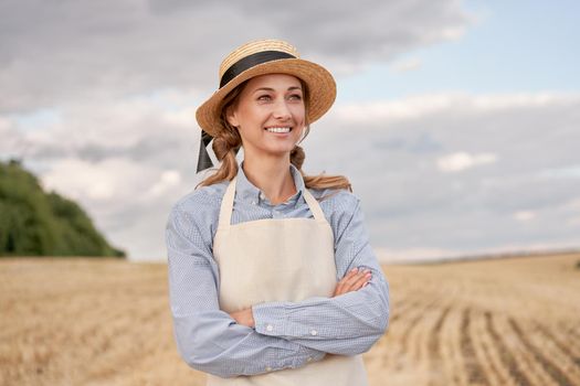 Woman farmer straw hat apron standing farmland smiling Female agronomist specialist farming agribusiness Happy positive caucasian worker agricultural field Girl arms crossed cloudy sky background