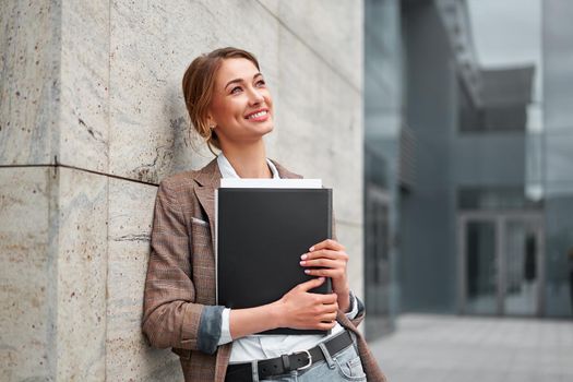 Cheerful business woman leaning at building office, holding documents, enjoying break outdoors