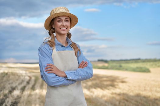 Woman farmer straw hat apron standing farmland smiling Female agronomist specialist farming agribusiness Happy positive caucasian worker agricultural field Girl arms crossed cloudy sky background