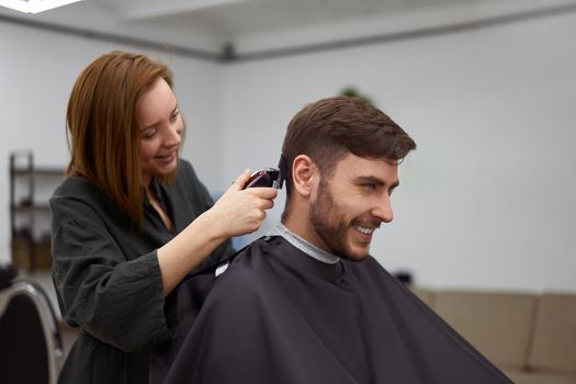 Stylish man sitting barber shop Hairstylist Hairdresser Woman cutting his hair Portrait handsome happy young bearded caucasian guy getting trendy haircut Attractive barber girl working serving client