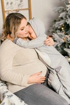 Mom plays with child. Happy family Portrait In Home pregnant mother embraces his little son. Happy new year. decorated Christmas tree Christmas morning bright living room Caucasian woman with child