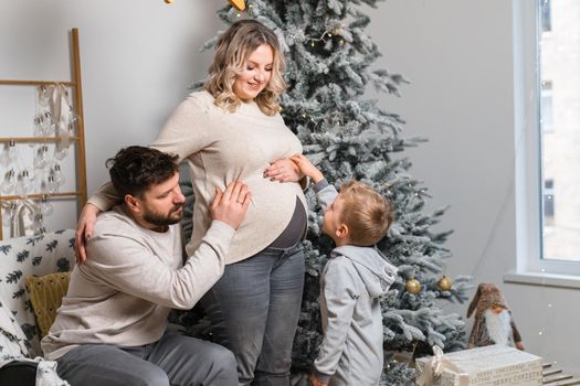 Christmas Family Happiness Portrait of dad, pregnant mom and little son sitting armchair at home near Christmas tree hug smile European young adult family holiday morning