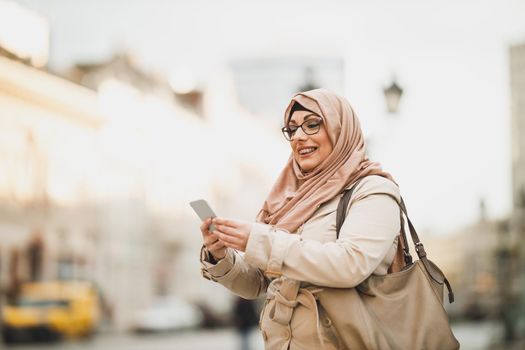 Smiling Muslim woman wearing a hijab and using her smartphone while standing in urban environment.