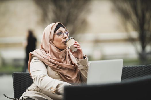 Smiling Muslim business woman wearing a hijab and using her laptop while sitting in urban cafe.