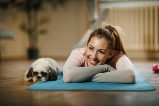 An attractive young woman lying on her yoga mat after a workout at home supporting by her pet dog.