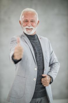Portrait of a smiling senior businessman standing in a modern office with thumb up and looking at camera. Selective focus.