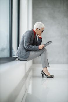 A confident mature businesswoman working on a digital tablet while sitting near the window in a modern office.