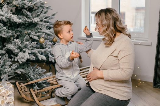 Holiday Christmas Caucasian mother playing with little cute son near decorated New year tree at home Family tradition