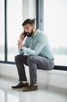 Portrait of a pensive businessman talking on a smart phone while sitting near the window in a modern office.