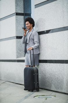 A successful black business woman standing with her luggage and talking voice message on a smart phone outside of the airport.